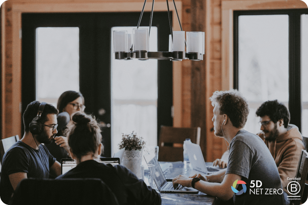 A group of five people work together at a table with laptops in a cosy, wooden room, embodying net zero values. Two certification logos, "5D Sustainability" and "Certified B Corporation," are visible in the lower right corner.