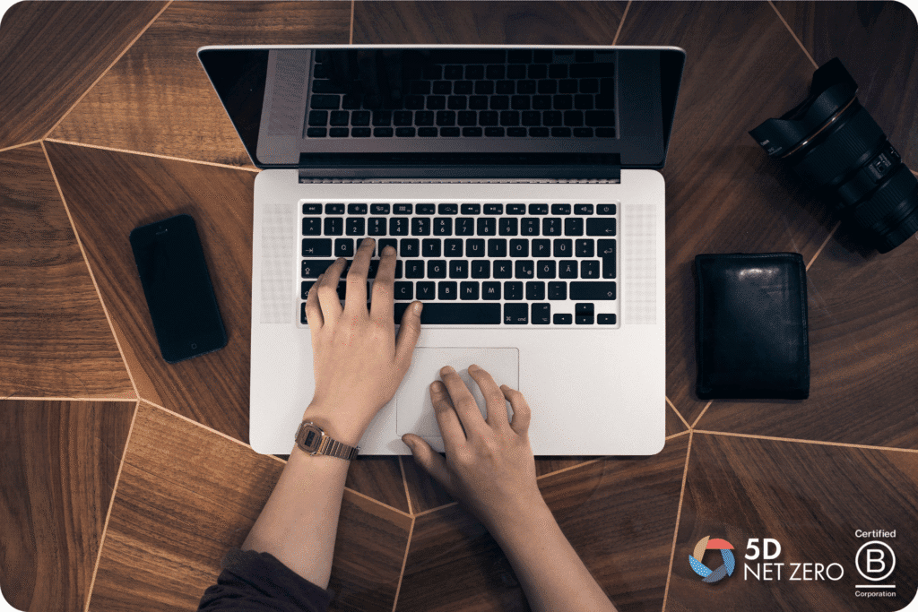 Overhead view of a person typing on a laptop at a geometric wooden table, with a mobile phone, camera, and wallet nearby. 5D Sustainability and Certified B Corporation logos highlight the company’s commitment to net zero initiatives in the bottom right corner.