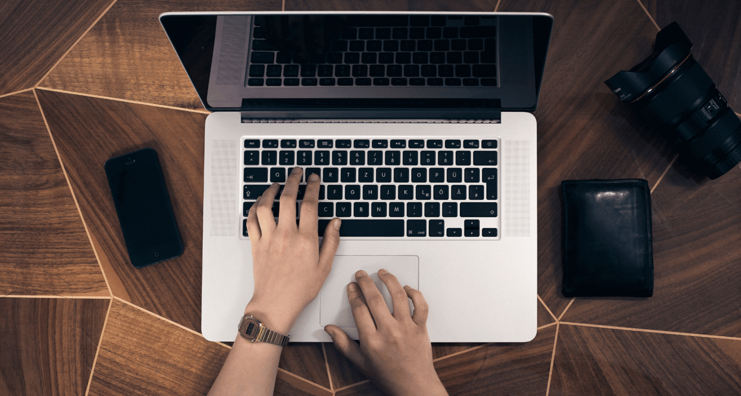 Overhead view of a person typing on a laptop at a geometric wooden table, with a mobile phone, camera, and wallet nearby. 5D Sustainability and Certified B Corporation logos highlight the company’s commitment to net zero initiatives in the bottom right corner.