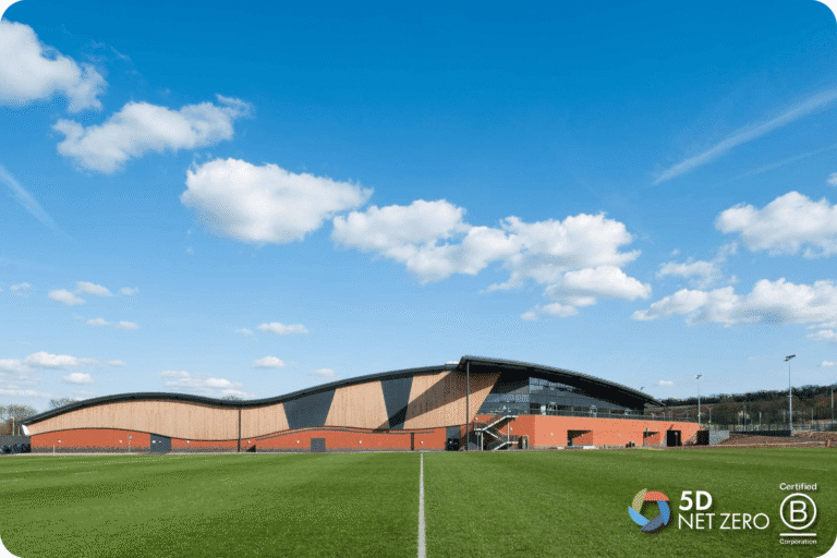 A modern, wave-shaped building with large glass windows sits beside a green sports pitch under a blue sky with scattered clouds—an inspiring space for business in Surrey. Logos for 5D Sustainability and Certified B Corporation appear in the bottom right corner.