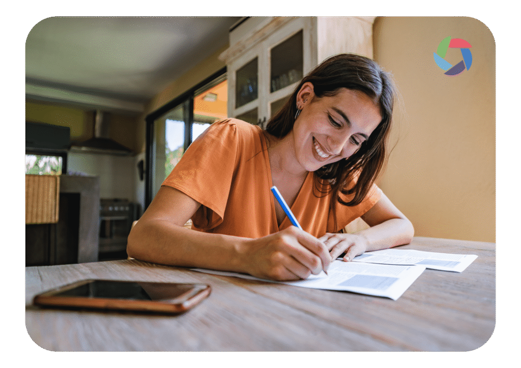 A young woman with long brown hair, wearing an orange shirt, sits at a table smiling as she writes in a notebook. A mobile phone lies on the table beside her. A colourful logo is in the top right corner.