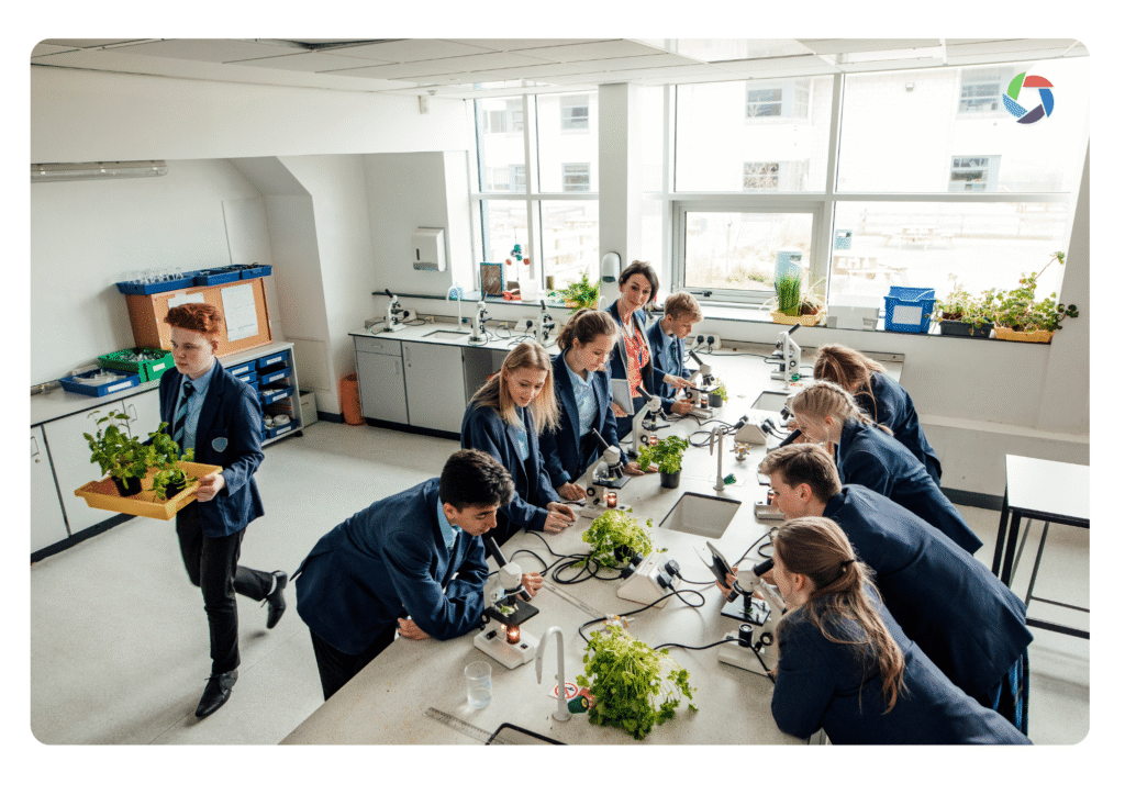Students in school uniforms work on science experiments with plants at lab tables in a bright classroom. One pupil carries a tray of plants, whilst others observe and use scientific equipment.