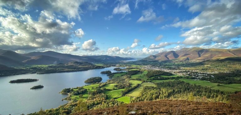 A scenic landscape features a large lake encircled by green fields, forests, and rolling hills under a blue sky with scattered clouds. A small town on the right nestles among the greenery, reflecting energy efficiency and carbon reduction efforts.