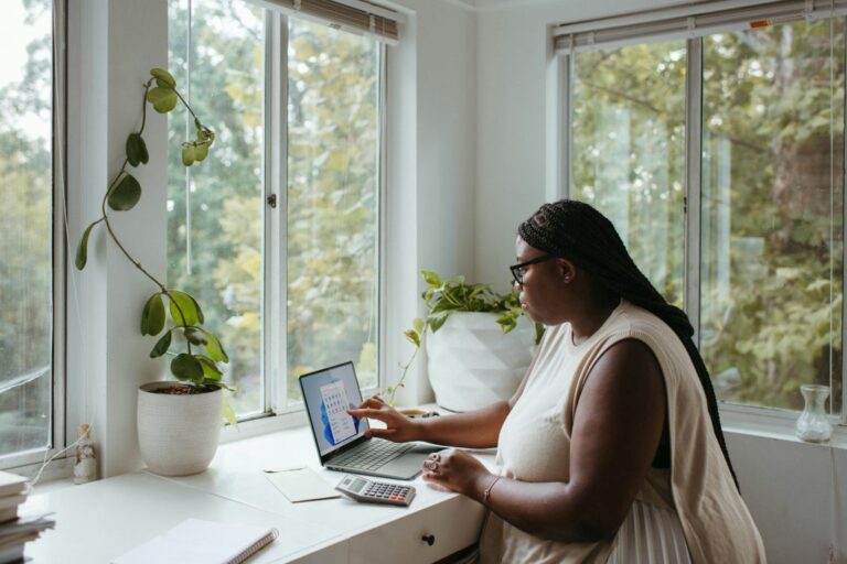 A woman sits at a desk by large windows with plants, using a laptop and calculator to analyse her business carbon footprint, appearing focused on the screen as natural light streams into the room.