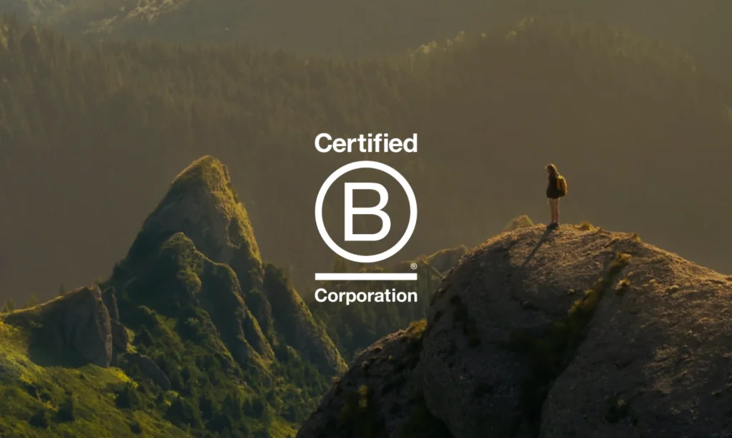 A person stands on a rocky mountain peak at sunrise with the Certified B Corporation logo and text overlaid in the centre.