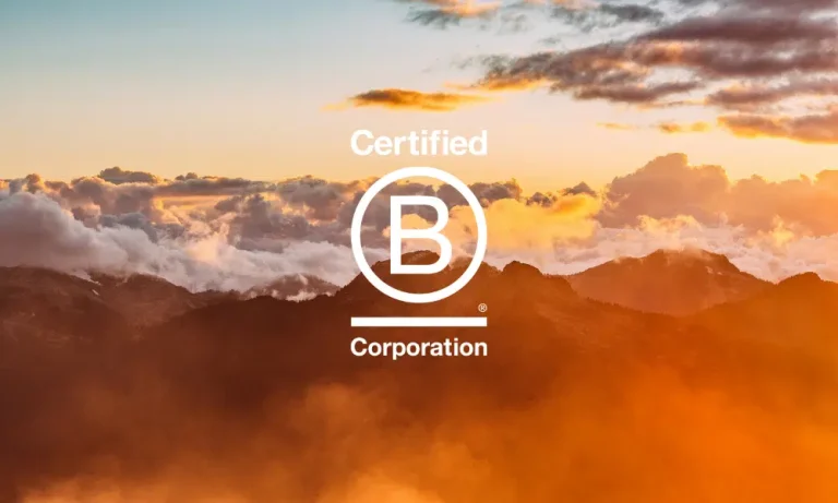 Golden sunlight illuminates mountain peaks and clouds at sunrise or sunset, creating a warm, glowing atmosphere above the rugged landscape with b-corp logo.