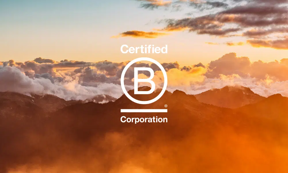 Golden sunlight illuminates mountain peaks and clouds at sunrise or sunset, creating a warm, glowing atmosphere above the rugged landscape with b-corp logo.