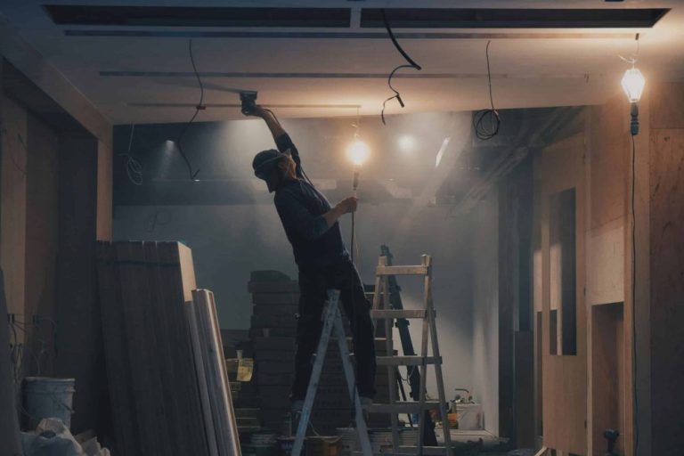 A person standing on a ladder works on the ceiling of a dimly lit, unfinished room with exposed wiring and building materials.