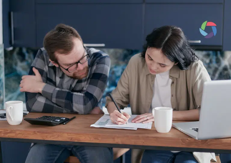 A man and woman sit at a table with papers, a laptop, calculator, and two mugs; the woman writes whilst the man looks on.