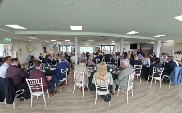 A group of people seated at round tables in a bright event room, listening to a presentation displayed on a screen at the front.