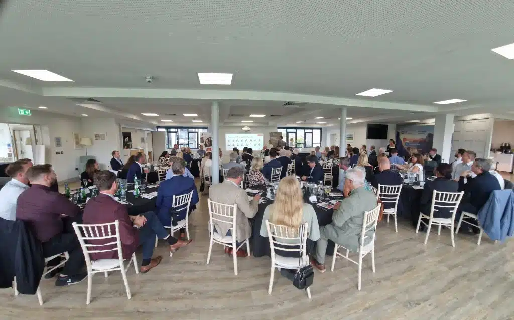 A group of people seated at round tables in a bright event room, listening to a presentation displayed on a screen at the front.