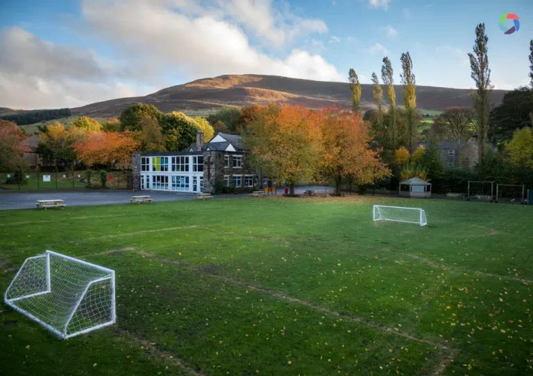 A grassy football pitch with two goals sits in front of a school building, surrounded by autumn trees and hills in the background under a partly cloudy sky.