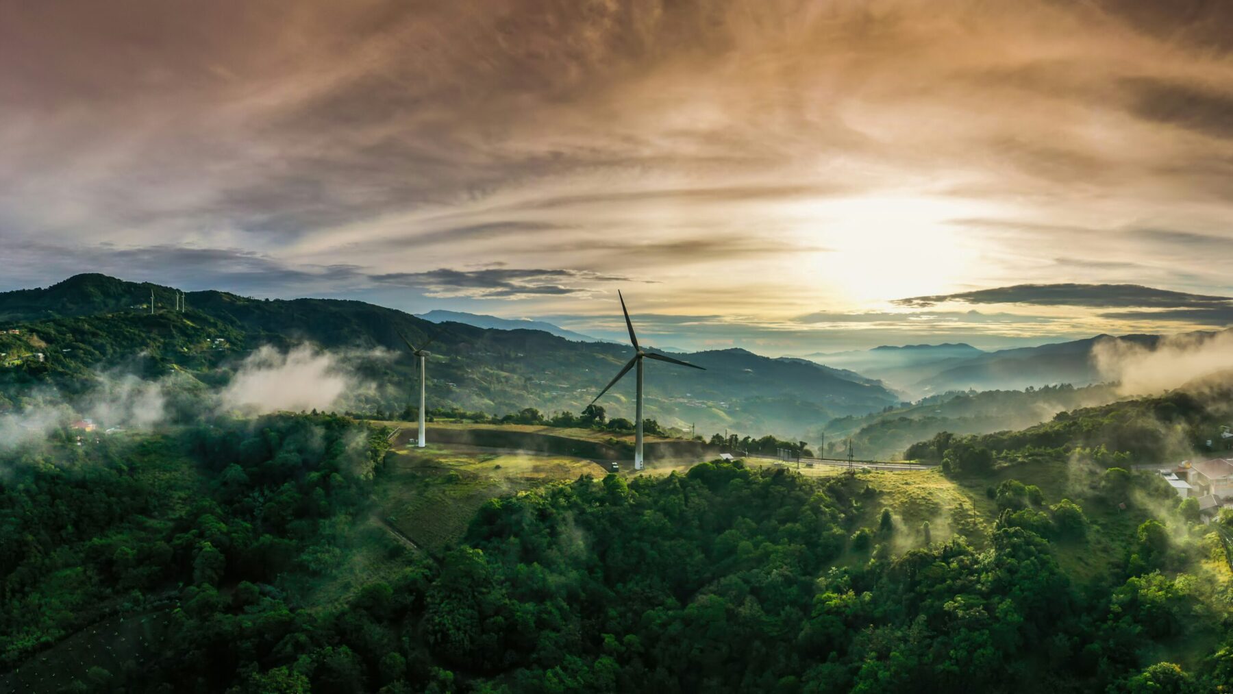 Wind turbines on a green hillside embody sustainability, rising through mist and distant mountains under a dramatic cloudy sky at sunrise or sunset.