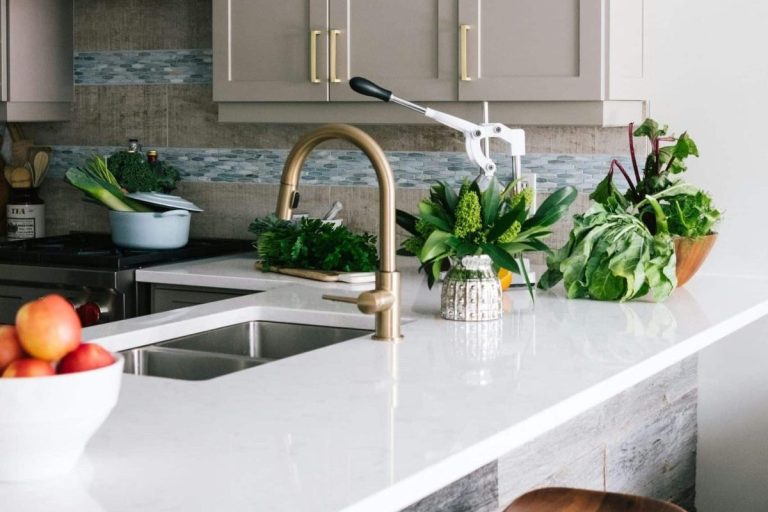 A modern kitchen with a white worktop, brass tap, fresh vegetables, a bowl of apples, and a pot on the hob.