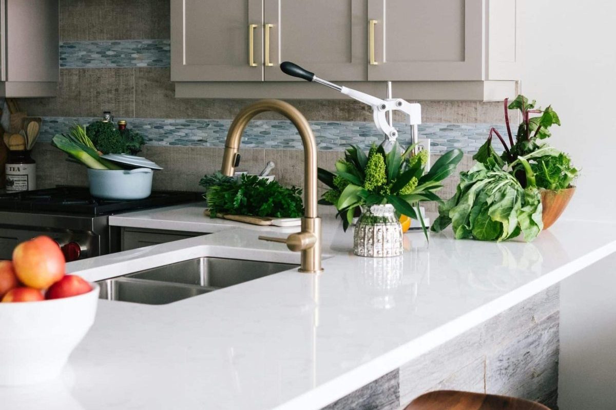 A modern kitchen with a white worktop, brass tap, fresh vegetables, a bowl of apples, and a pot on the hob.