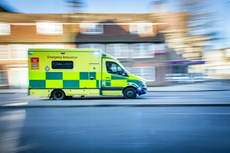 A yellow and green emergency ambulance drives quickly along a city street, with buildings blurred in the background to indicate movement.