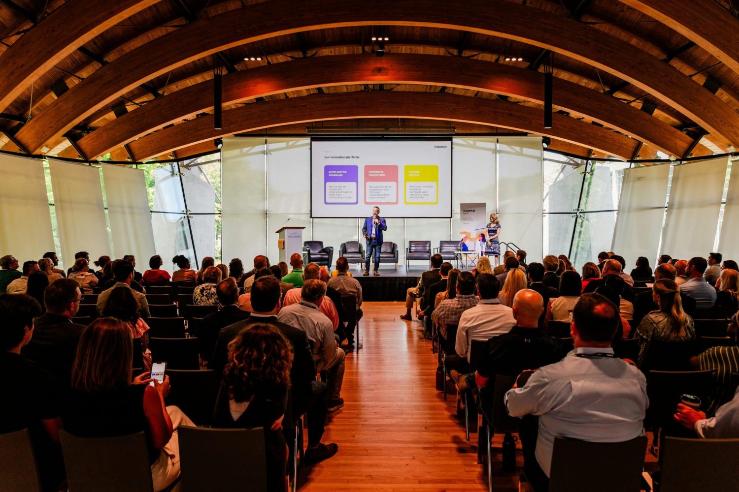 A speaker presents on stage to an audience in a modern conference hall with a large screen displaying a slide of four coloured boxes.