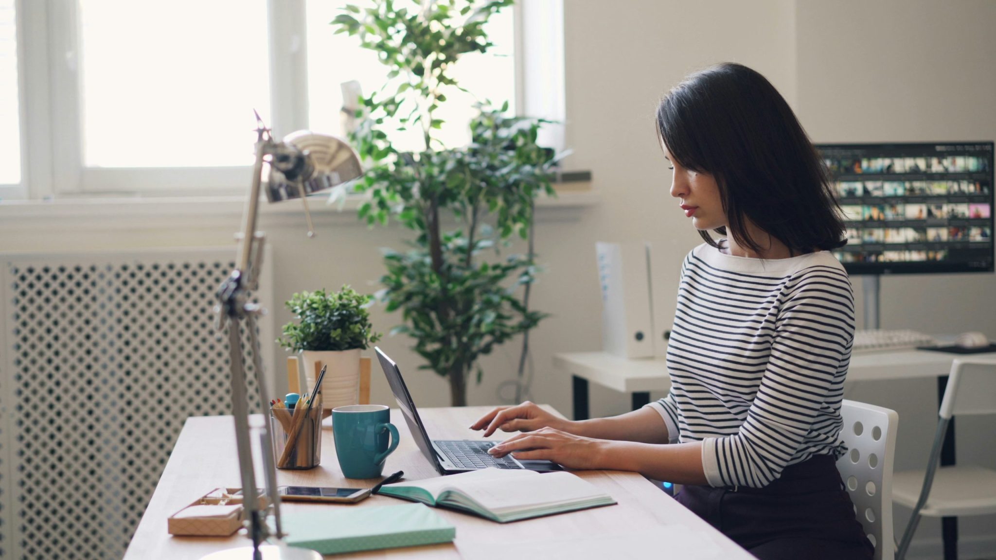 Woman sitting at a desk in a bright office, typing on a laptop with a notebook, mug, and plants nearby. Computer monitor and office supplies are visible in the background.
